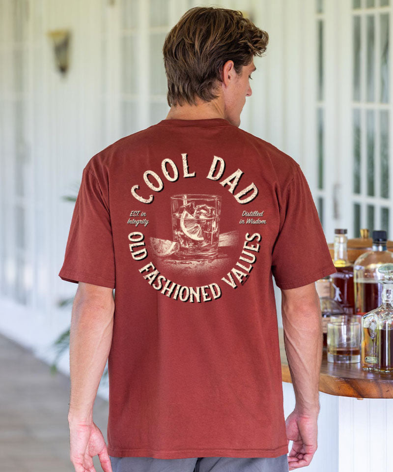 Man wearing a brown t-shirt with 'Cool Dad' text and graphic, standing in a room with bottles in the background. View 2