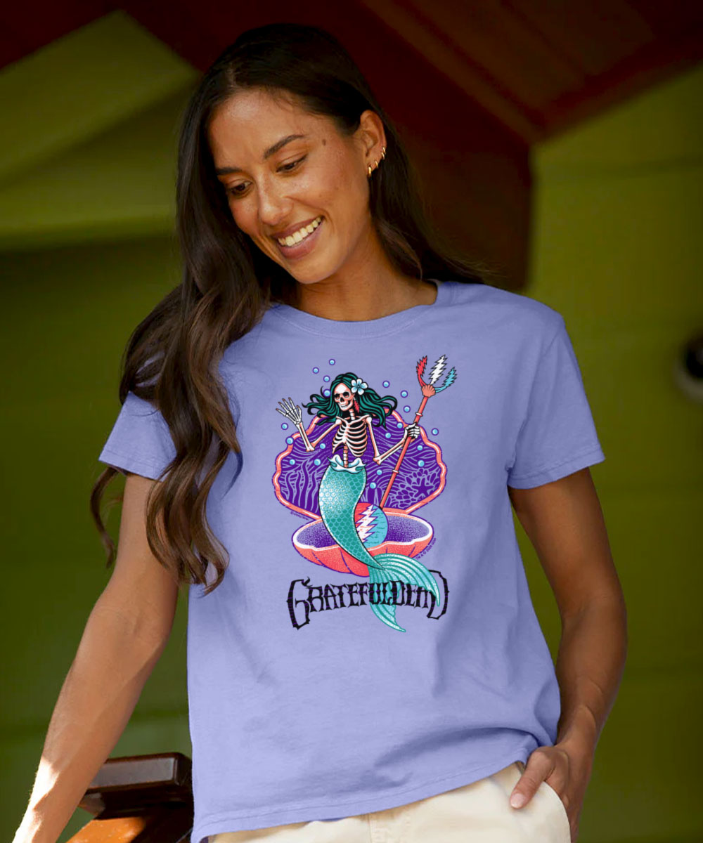 A woman wearing a light periwinkle blue t-shirt with a colorful graphic of a skeleton mermaid and the Grateful Dead logo. The background is a soft-focus olive green wall. View 2
