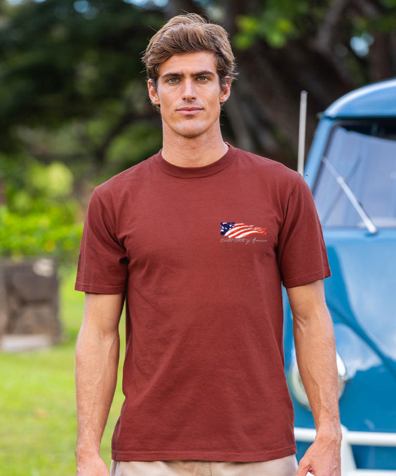 Man wearing a brown t-shirt with 'Hawaii' print, standing outdoors with greenery and a vintage car in the background. View 3