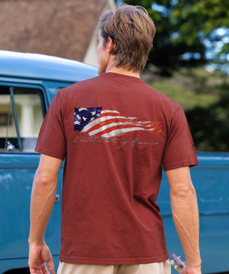 Man wearing a brown t-shirt with an American flag design, standing in front of a blue truck. View 2