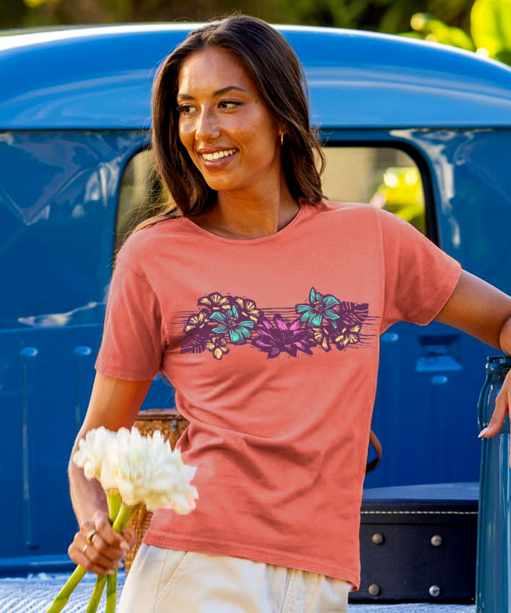 Woman in a coral t-shirt with floral design holding flowers, standing next to a blue truck. View 2