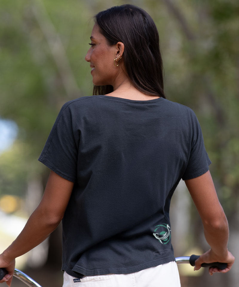 Woman wearing a black t-shirt with a logo, standing outdoors with blurred greenery in the background. View 3