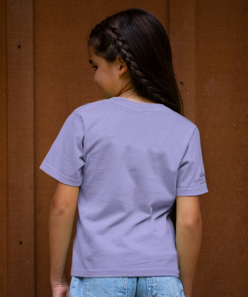 Person wearing a light purple t-shirt with a logo on the back, standing against a brown wall. View 3