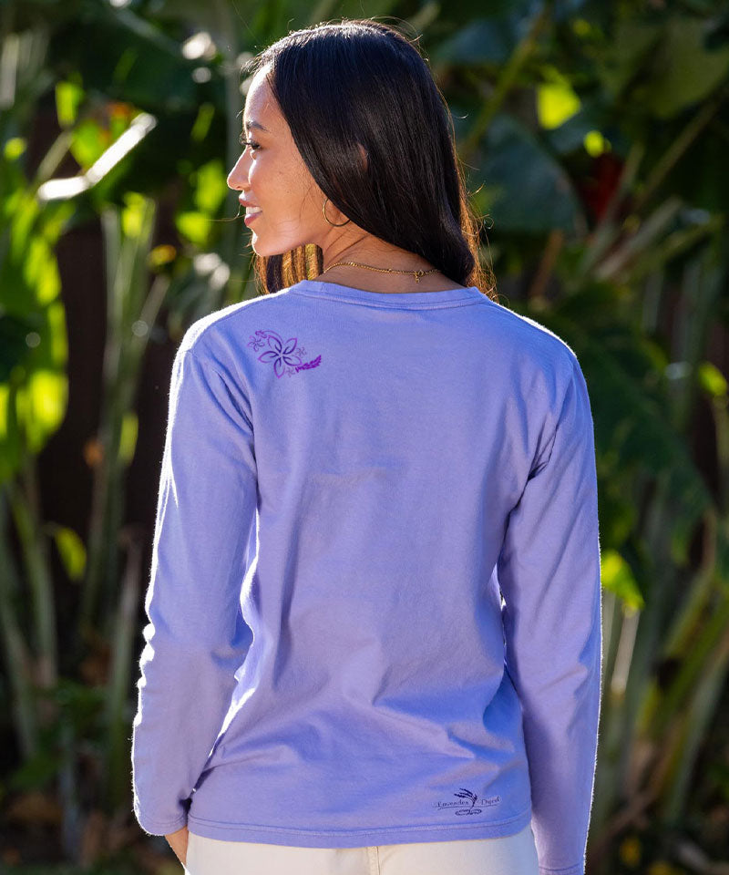 Woman wearing a light blue long-sleeve shirt with a logo on the back, standing in front of green foliage. View 3