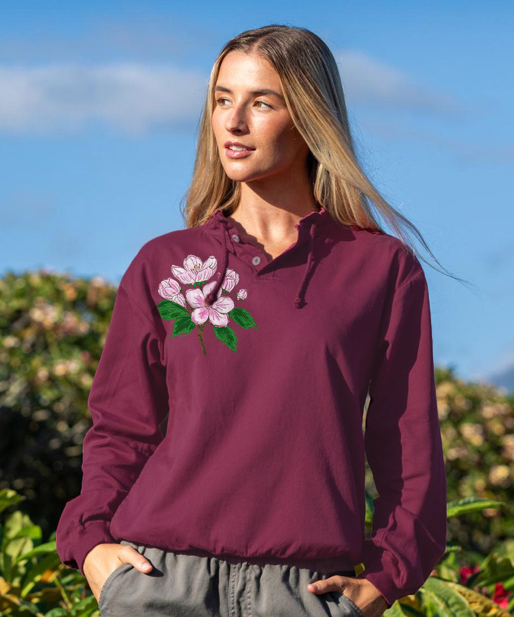 A woman wearing a maroon hooded pullover sweatshirt with a pink and green floral embroidery on the left chest, set against an outdoor background of green foliage and a blue sky.