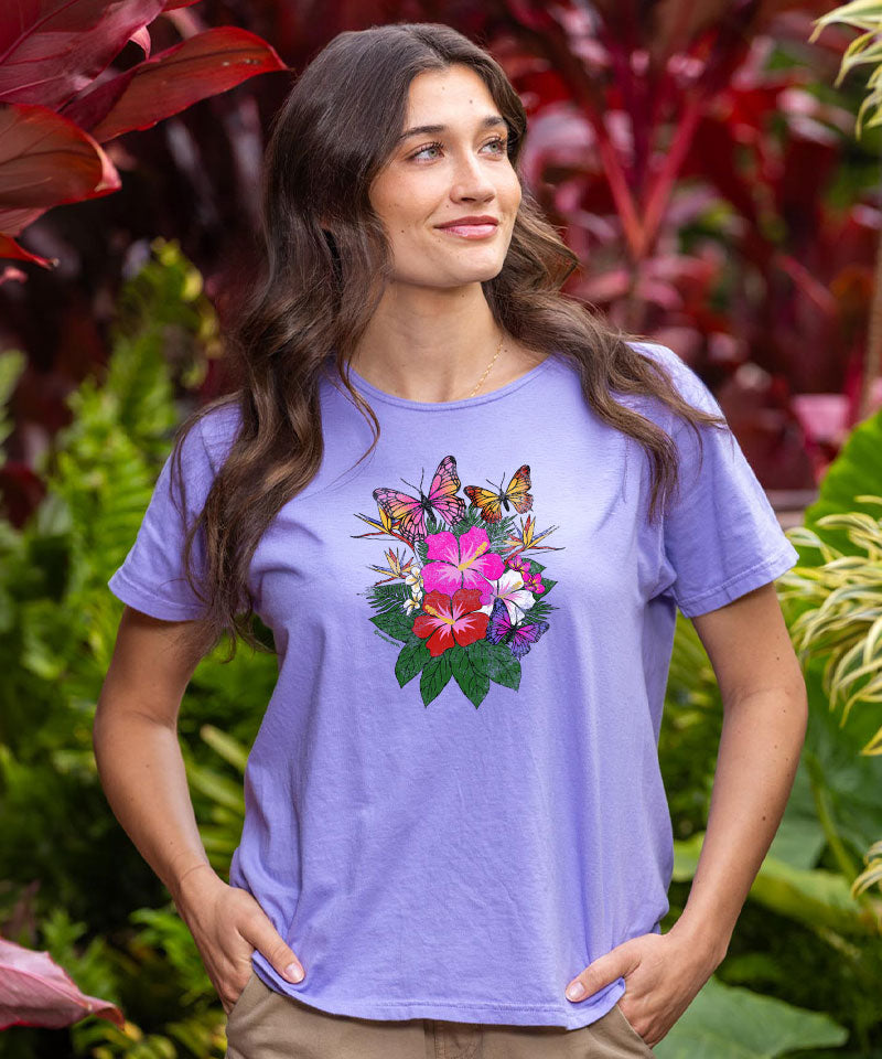 Woman wearing a purple t-shirt with a floral and butterfly design, standing in front of green foliage. View 2
