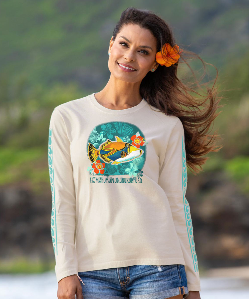 A woman wearing a cream-colored long-sleeve t-shirt with a colorful graphic of a yellow fish on a teal background and turquoise patterned sleeves, set against a blurred outdoor beach backdrop.