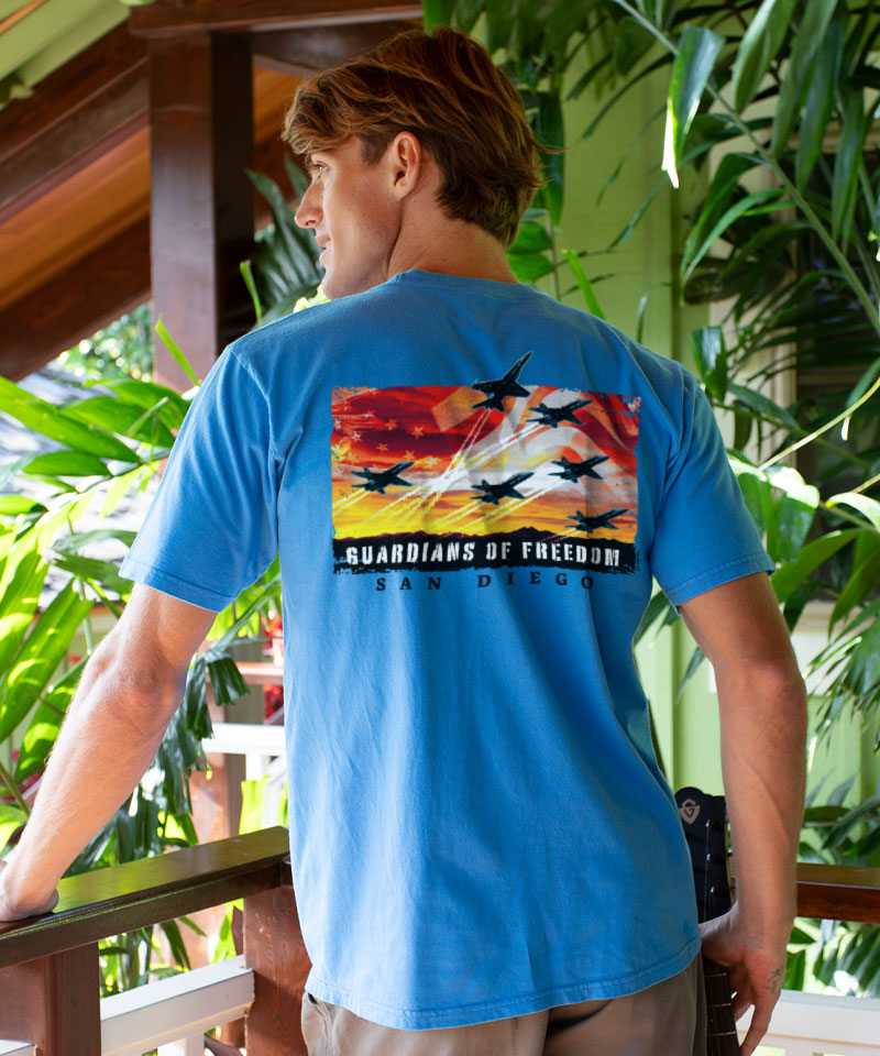 Man wearing a blue t-shirt with a graphic design and text, standing outdoors with greenery in the background. View 2