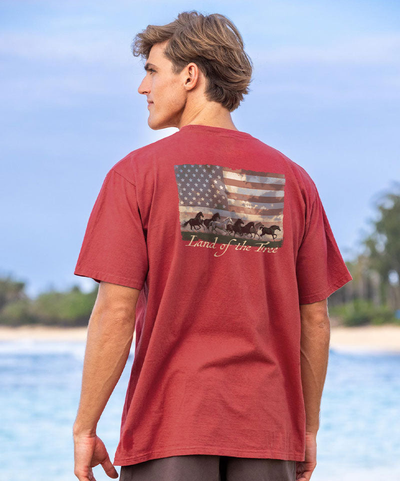 Person wearing a red t-shirt with a graphic design of an American flag and text, standing by a beach. View 2