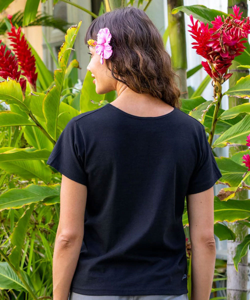 A woman viewed from behind wearing a plain black short-sleeve t-shirt, standing outdoors in front of lush green tropical plants and bright red flowers. She has a pink flower in her hair. View 3