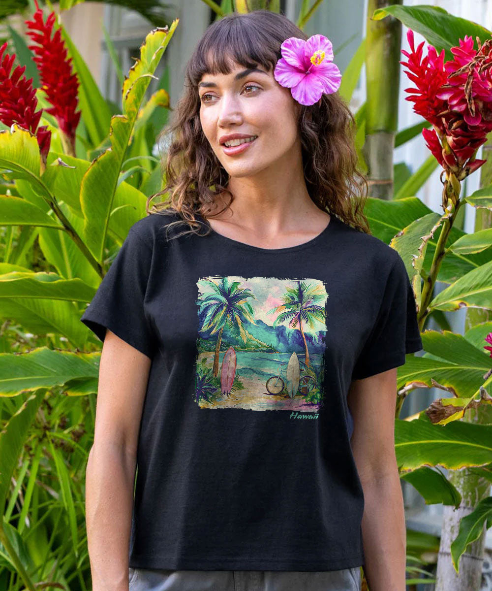 A woman wearing a black short-sleeve graphic t-shirt with a tropical beach print, standing outdoors in front of lush green tropical foliage and red flowers. She has a pink hibiscus flower in her hair. View 2