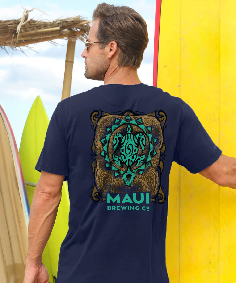 A man wearing a navy blue t-shirt with a large green and gold tribal design and the text 'MAUI BREWING CO' on the back, standing in front of a bright yellow wall with surfboards in the background. View 2