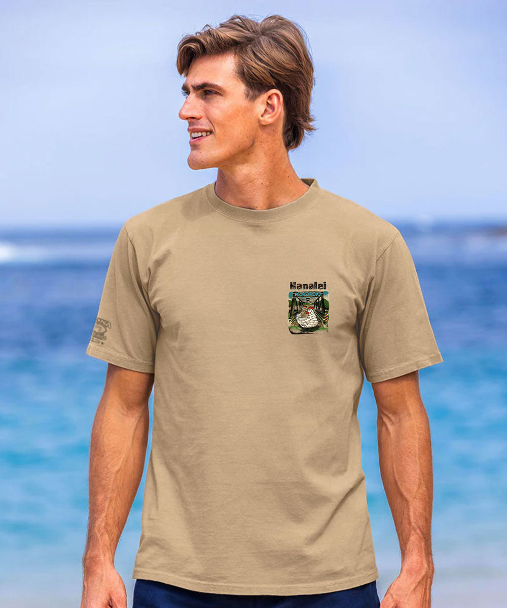 A man wearing a tan crewneck t-shirt with a small 'Hanalei' tropical graphic on the left chest, standing against a blurred beach and ocean background. View 3