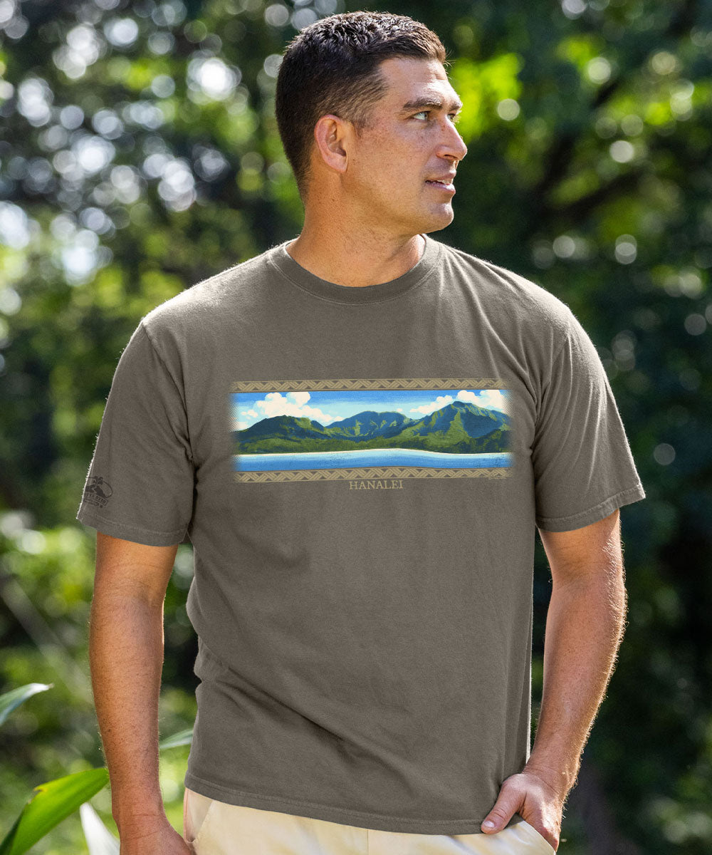 A man wearing a taupe brown short-sleeve t-shirt with a scenic mountain landscape graphic on the chest, set against a blurred green outdoor background. View 2