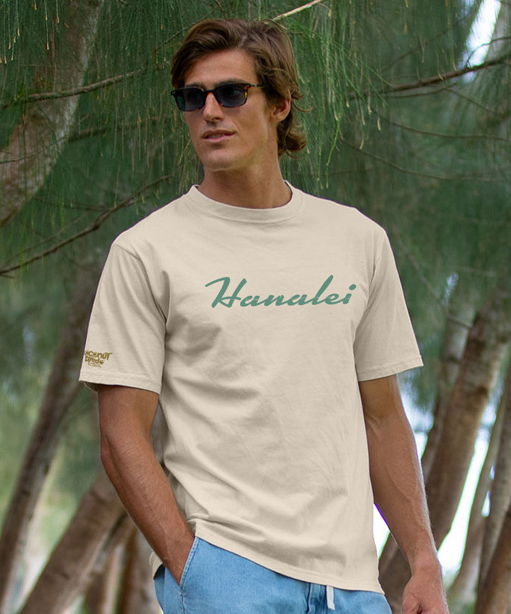 A man wearing a light beige crewneck t-shirt with 'Hanalei' printed in green script across the chest, set against a background of pine trees. View 3