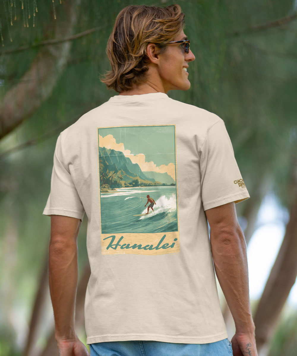 A man wearing a beige t-shirt with a large retro-style graphic print of a surfer in Hanalei, Hawaii, photographed outdoors against a blurred green background. View 2