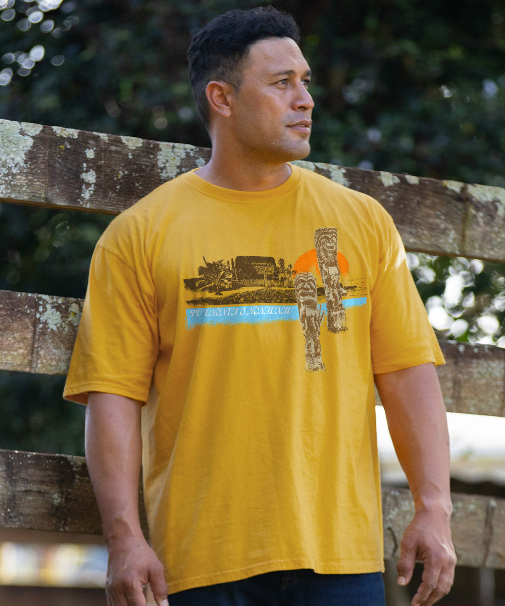 A man wearing a mustard yellow graphic t-shirt with a Hawaiian-themed print, standing outdoors in front of a weathered wooden fence. View 2