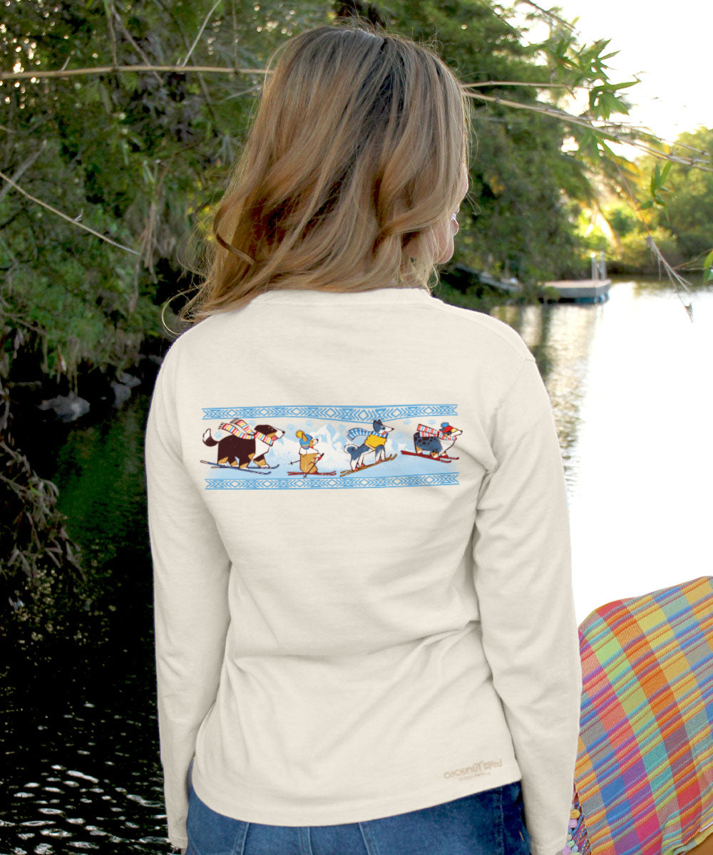 Woman wearing a white long-sleeve shirt with a graphic of dogs skiing on the back, standing by a body of water. View 3