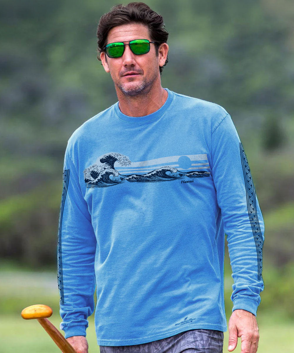 A man wearing a light blue long-sleeve graphic shirt with a wave print on the chest, green-tinted sunglasses, and holding a wooden paddle, set against a blurred green outdoor background. View 2