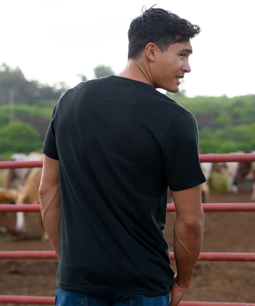 A man wearing a black short-sleeve crewneck t-shirt, shown from the back in an outdoor setting with a red metal fence and blurred greenery in the background. View 3