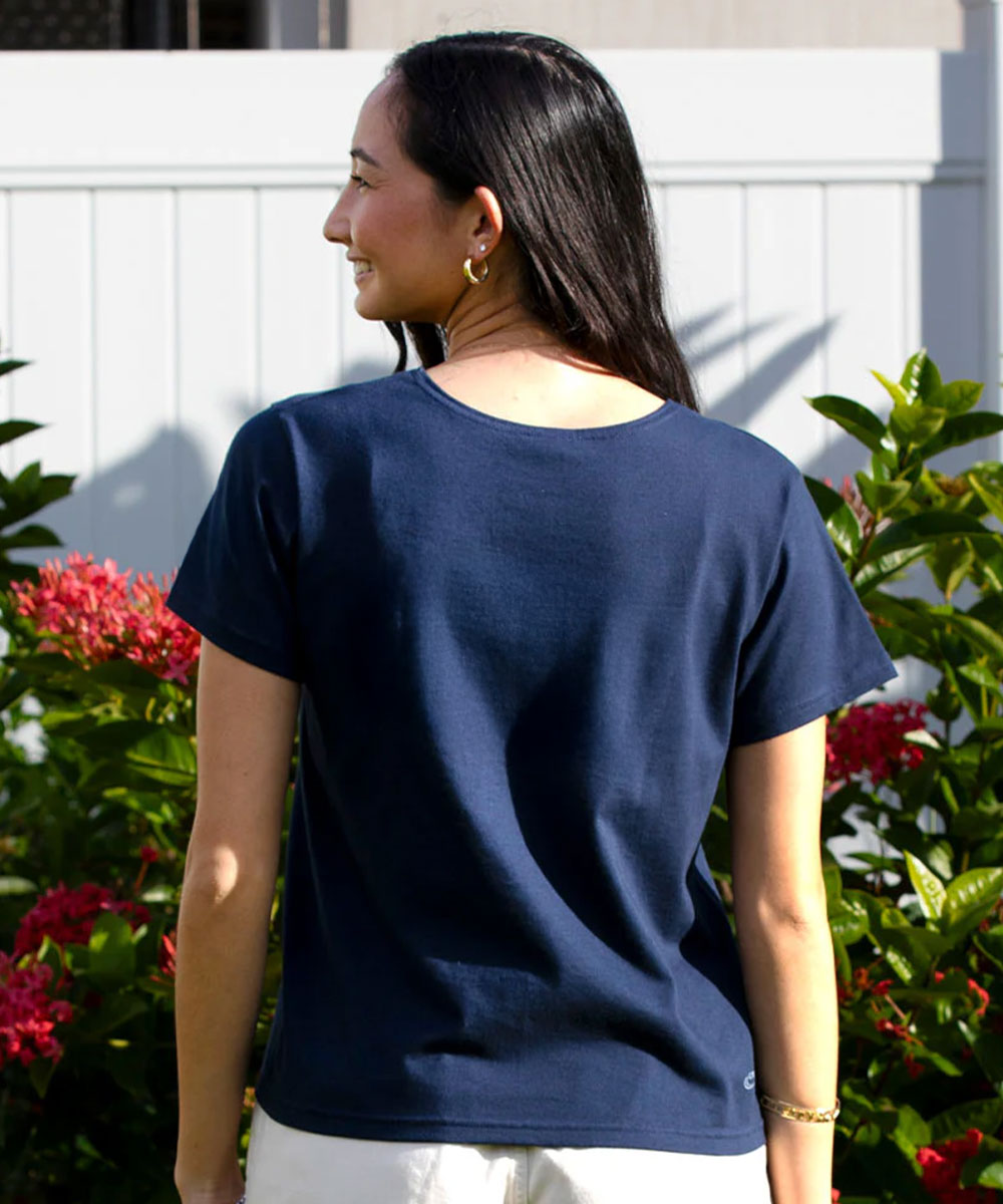 A woman shown from the back wearing a navy blue short-sleeve crewneck t-shirt, standing outdoors in front of a white fence and green plants with red flowers. View 3