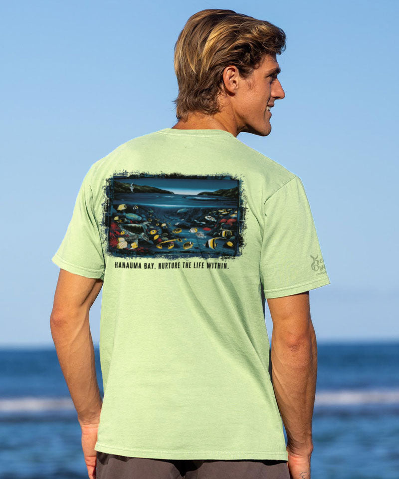 Man wearing a light green t-shirt with a graphic design on the back, standing by the ocean.