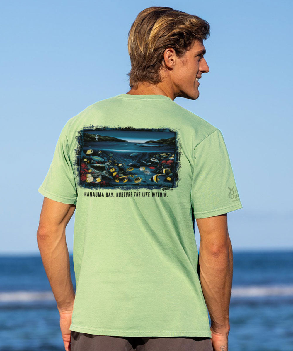 Man wearing a light green t-shirt with a graphic design on the back, standing by the ocean. View 2