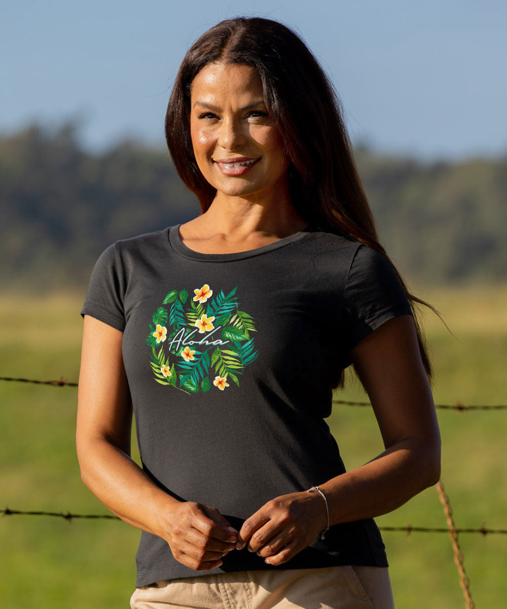 A woman wearing a dark charcoal gray t-shirt with a green tropical floral wreath graphic and the word 'Alahea' written in white script, standing outdoors in a grassy field. View 2