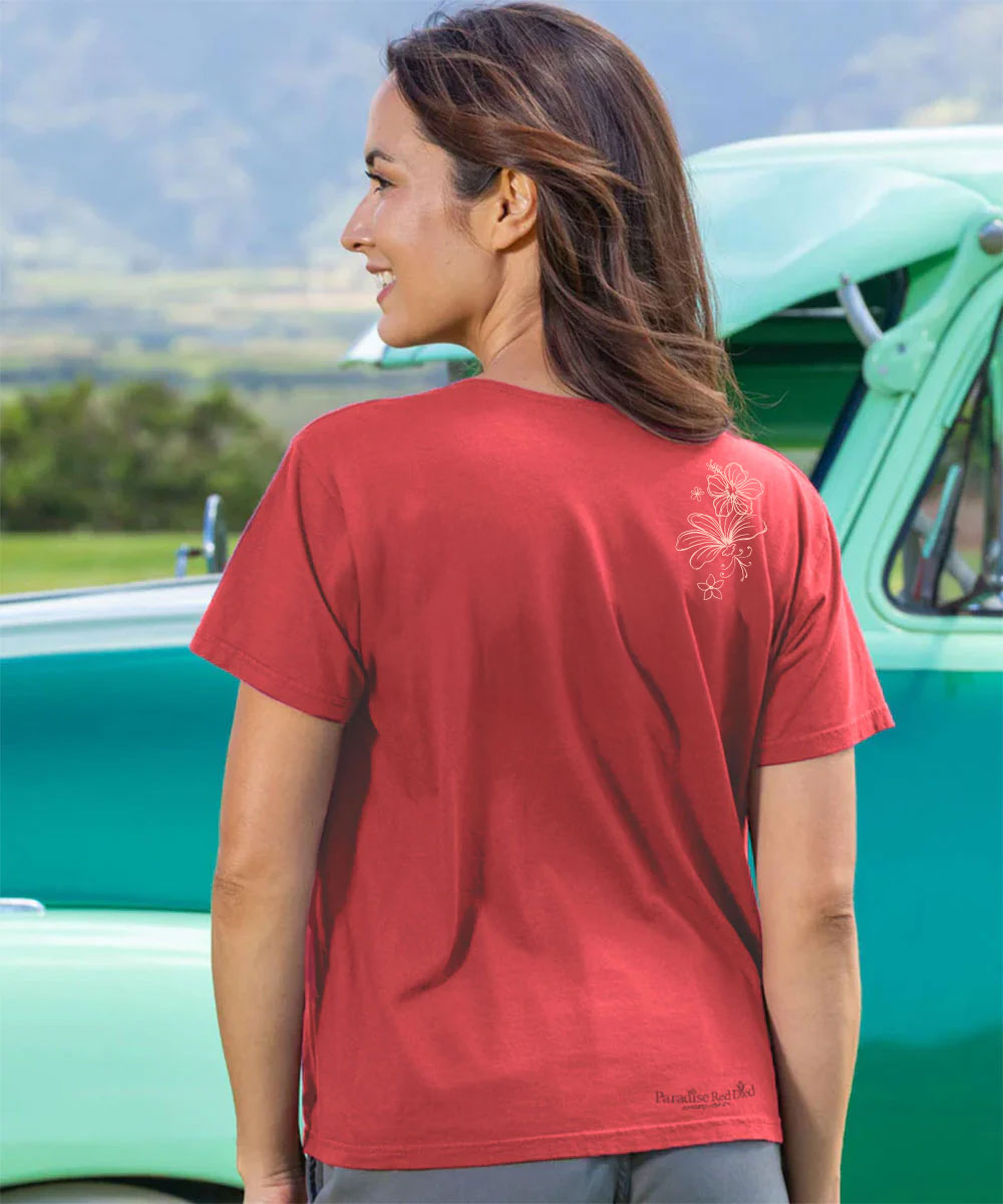 A woman shown from the back wearing a red short-sleeve t-shirt with a small white floral graphic on the upper right back and a small 'Pineapple Patch' brand logo near the bottom left hem. She is standing outdoors in front of a vintage mint green truck with a blurred mountain landscape in the background. View 3