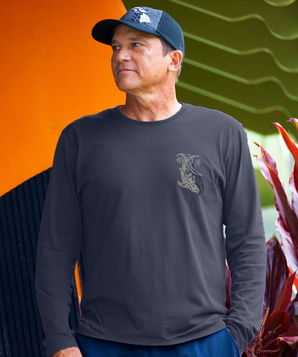 A man wearing a dark charcoal gray long-sleeve t-shirt with a small embroidered graphic on the left chest, paired with a navy blue baseball cap and blue pants, set against a vibrant orange and green background. View 3