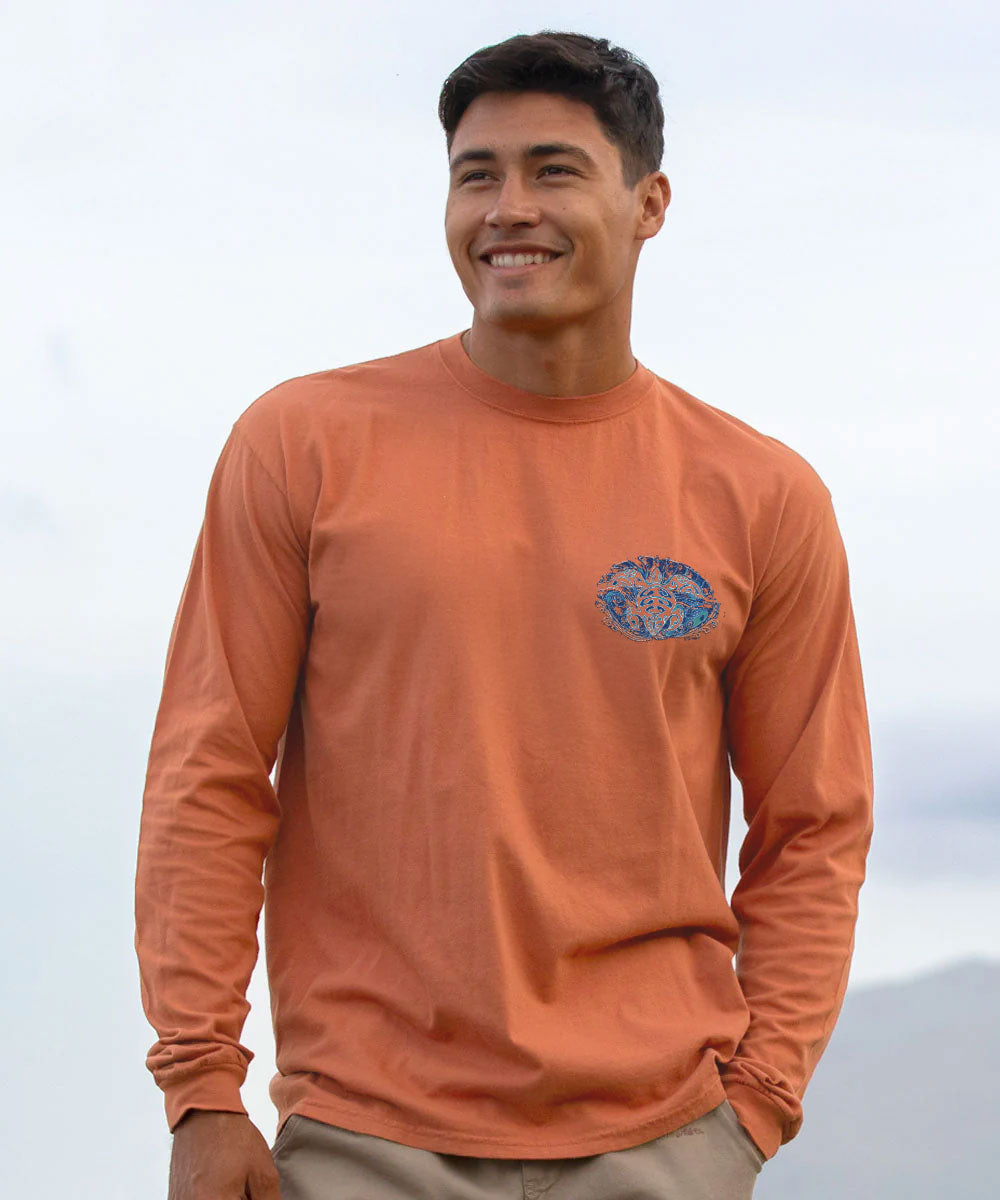 A smiling man wearing a long-sleeve crewneck shirt in a muted orange color, featuring a blue and white graphic logo on the left chest, set against a bright, overcast sky. View 3