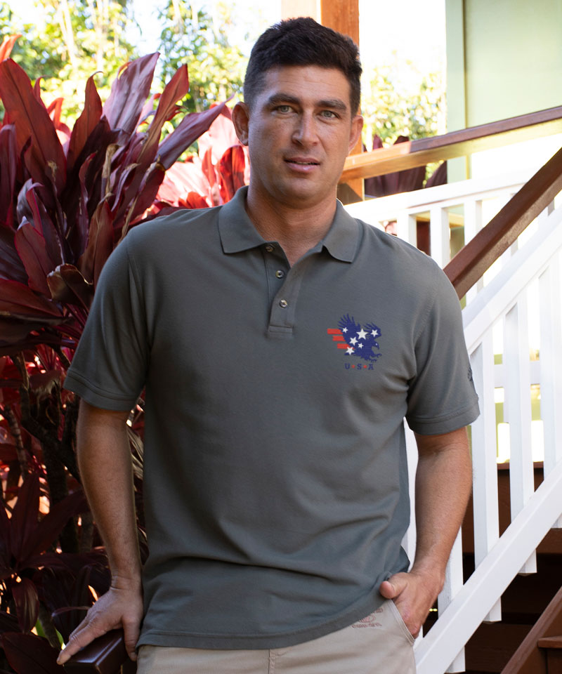 Man wearing a gray polo shirt with a logo, standing outdoors near plants and a staircase. View 2
