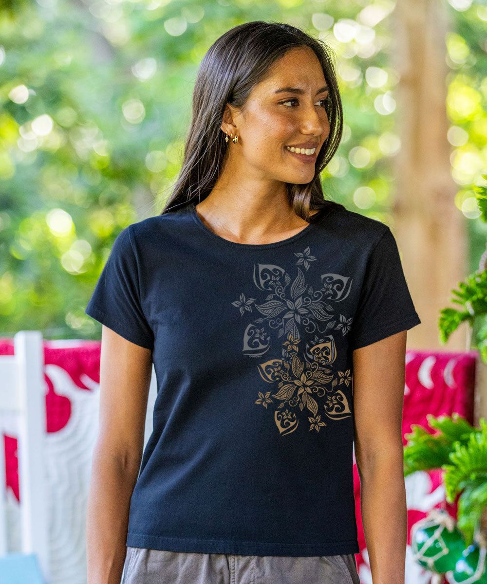 A woman wearing a dark navy blue t-shirt with a gold-toned floral mandala graphic on the front, set against a blurred green outdoor background. View 2