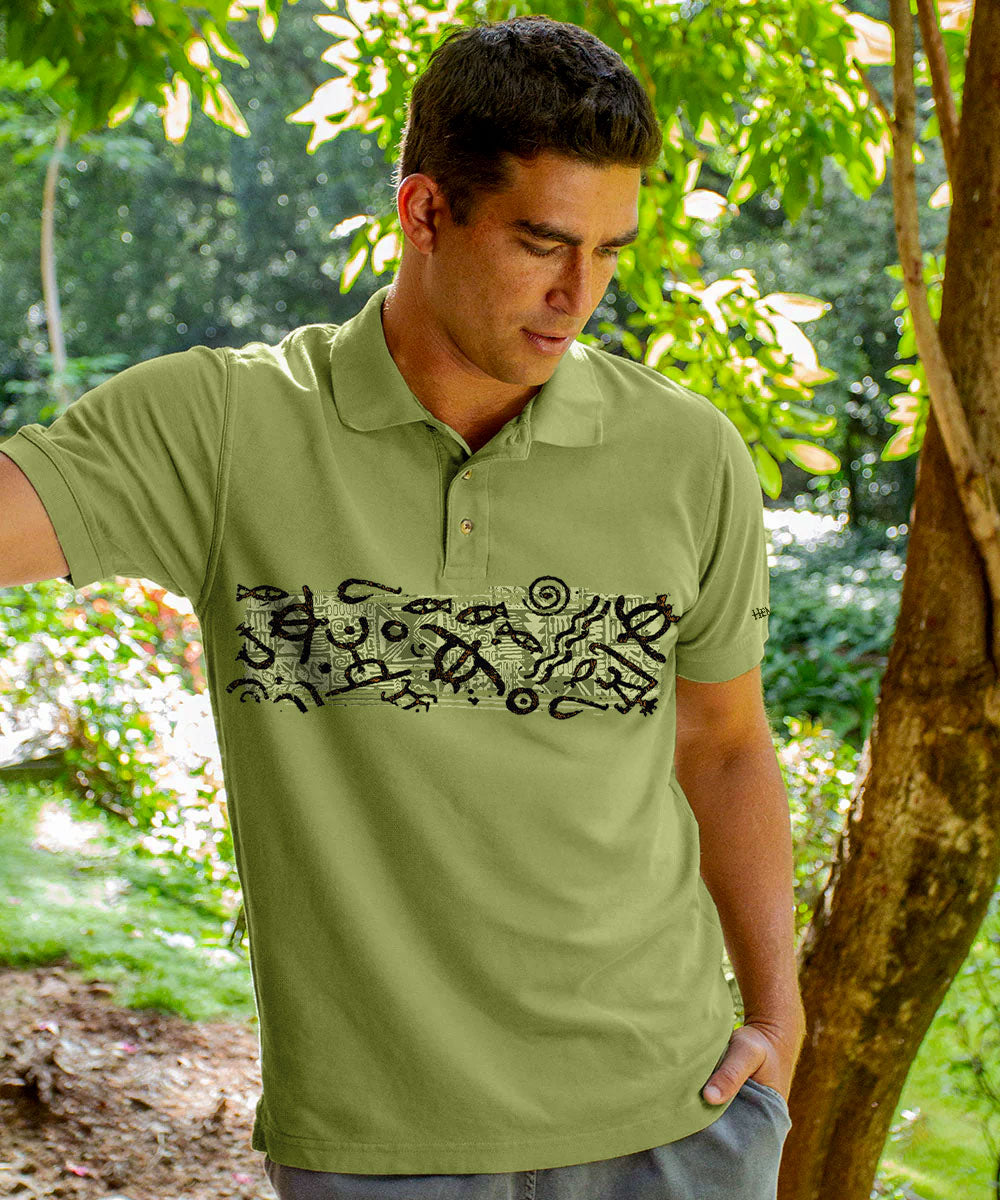 A man wearing an olive green short-sleeve polo shirt with a black abstract graphic across the chest, standing outdoors in a wooded setting. View 2