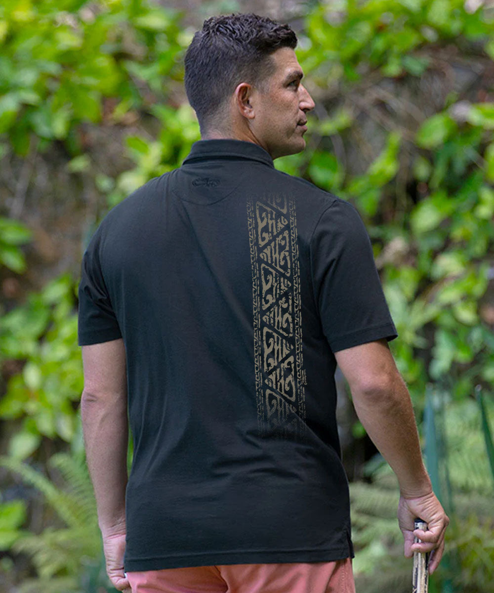 A man wearing a black short-sleeve polo shirt with a vertical geometric tribal-style pattern on the back, photographed outdoors in front of lush green tropical foliage. View 3