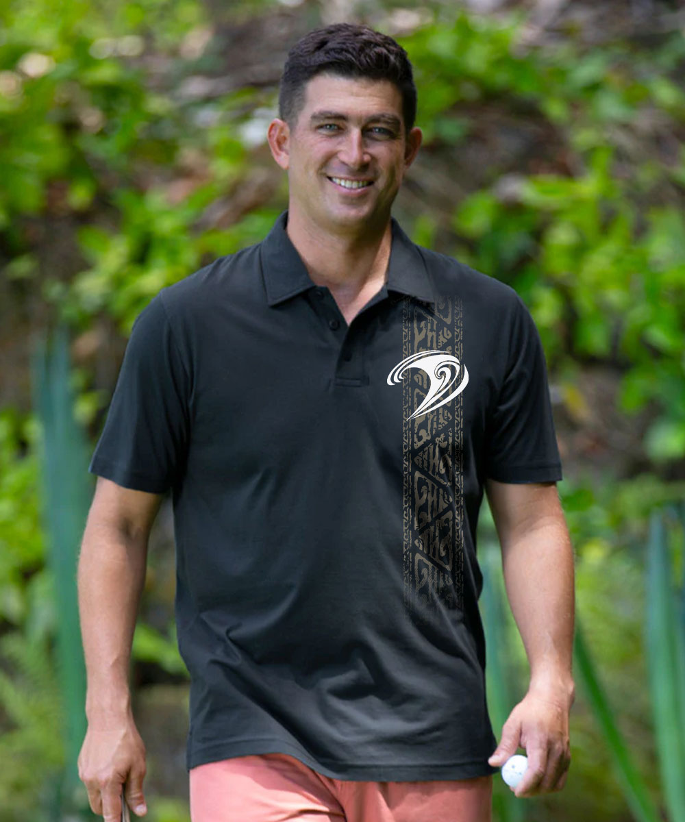 A man wearing a black short-sleeve polo shirt with a white and black Polynesian-style wave graphic on the chest, set against a lush green tropical background. View 2