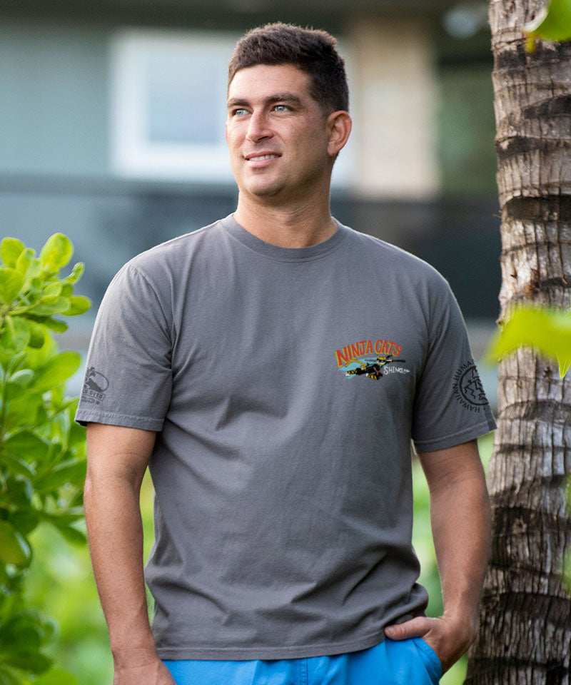 Man wearing a gray t-shirt with a logo, standing outdoors with greenery in the background View 3