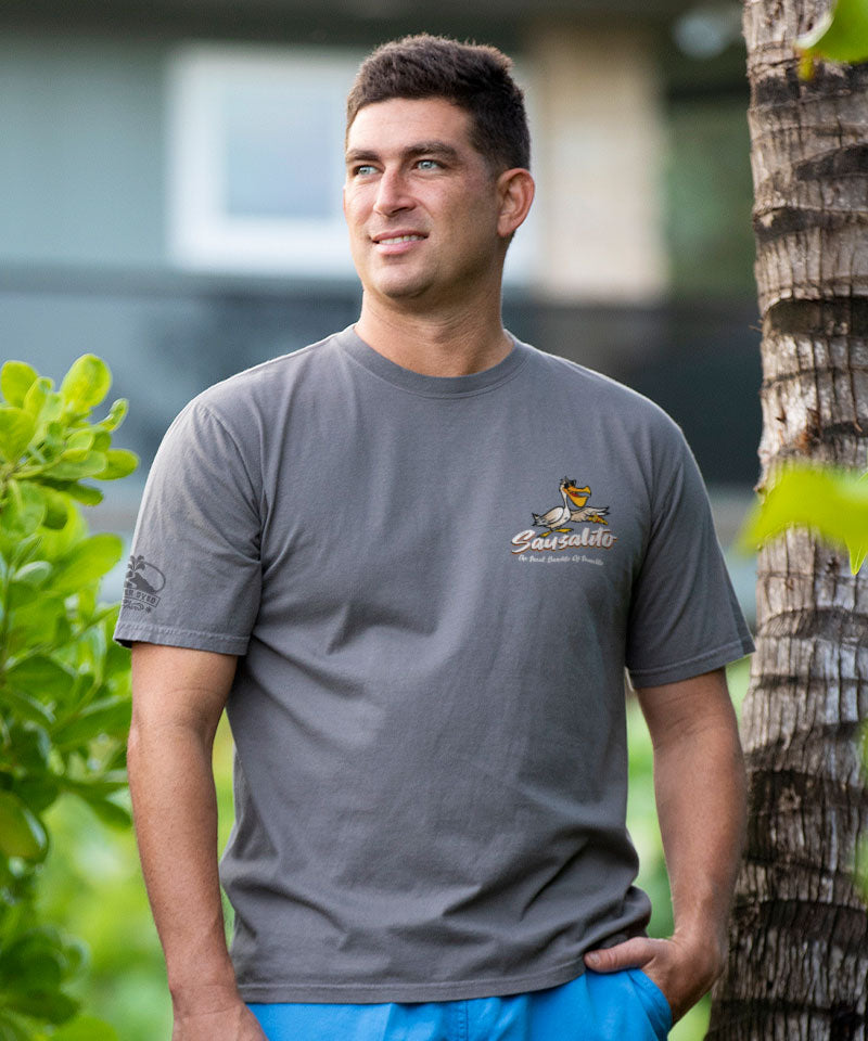Man wearing a gray t-shirt with a colorful graphic on the front, standing outdoors with greenery and a tree in the background. View 3
