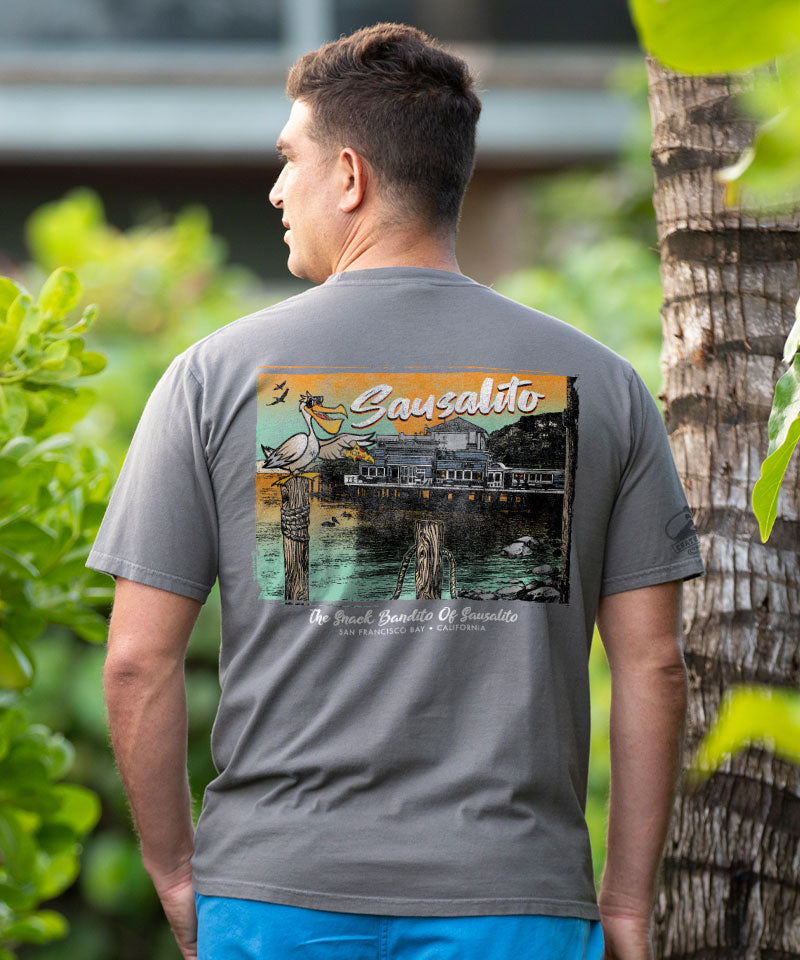 Man wearing a gray t-shirt with a colorful graphic on the back, standing outdoors with greenery and a tree in the background. View 2