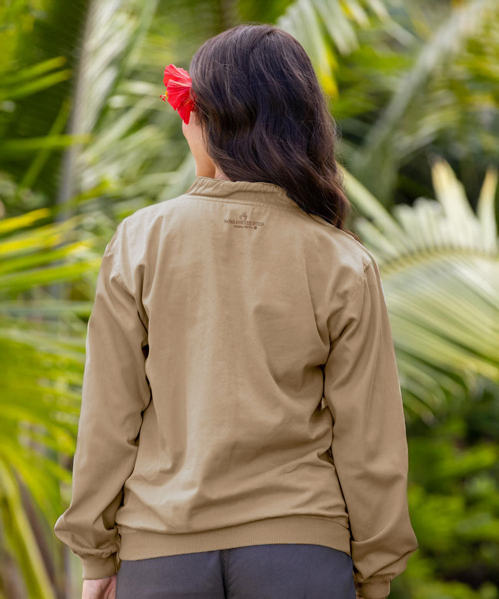 A woman viewed from behind wearing a light beige long-sleeve zip-up jacket, with a red flower in her hair, set against a background of green tropical palm leaves. View 3
