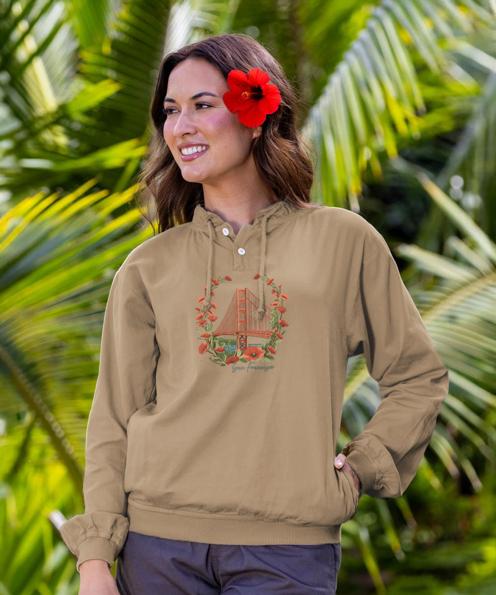 A woman wearing a light taupe pullover hoodie with a Golden Gate Bridge graphic on the chest, set against a lush green tropical background with palm leaves. View 2