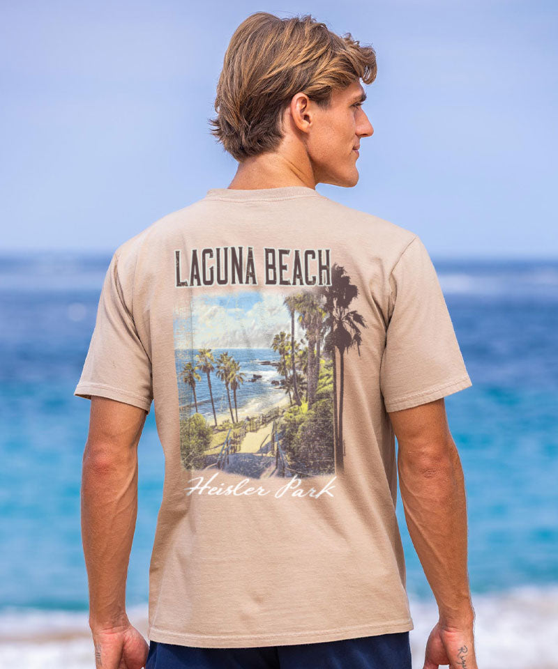 Person wearing a beige t-shirt with 'Laguna Beach' and palm tree design, standing on a beach. View 2
