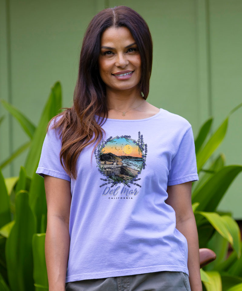 Woman wearing a purple t-shirt with a scenic graphic, standing outdoors against a green wall. View 2