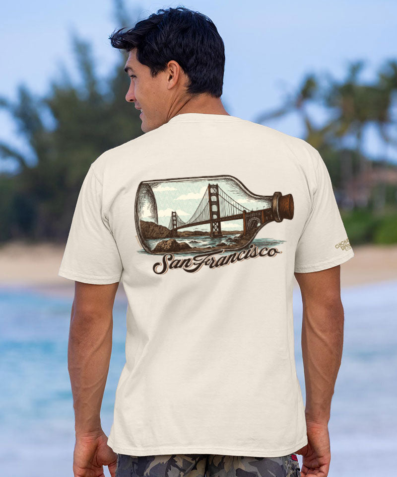 Man wearing a t-shirt with a graphic design of the Golden Gate Bridge in a bottle, standing on a beach. View 2