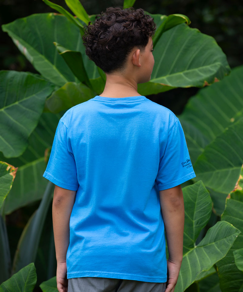 Person wearing a blue t-shirt with a brand logo, standing in front of green foliage View 3