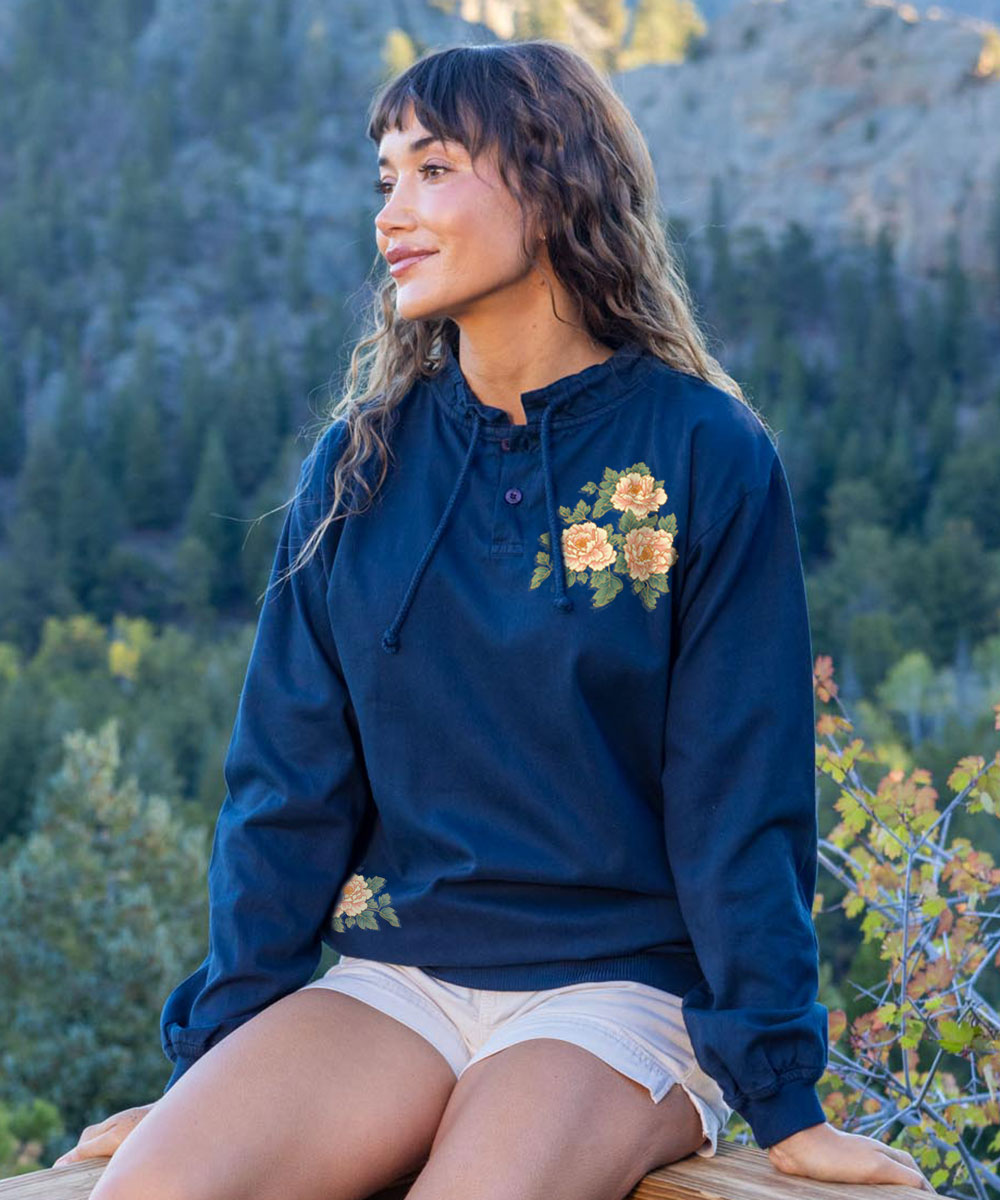 A woman wearing a navy blue hooded sweatshirt with yellow and orange floral embroidery on the chest, sitting outdoors in front of a forested mountain background.