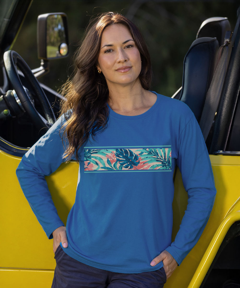 A woman wearing a bright blue long-sleeve crewneck shirt with a tropical floral and leaf pattern horizontal band across the chest, standing next to a yellow vehicle. View 2