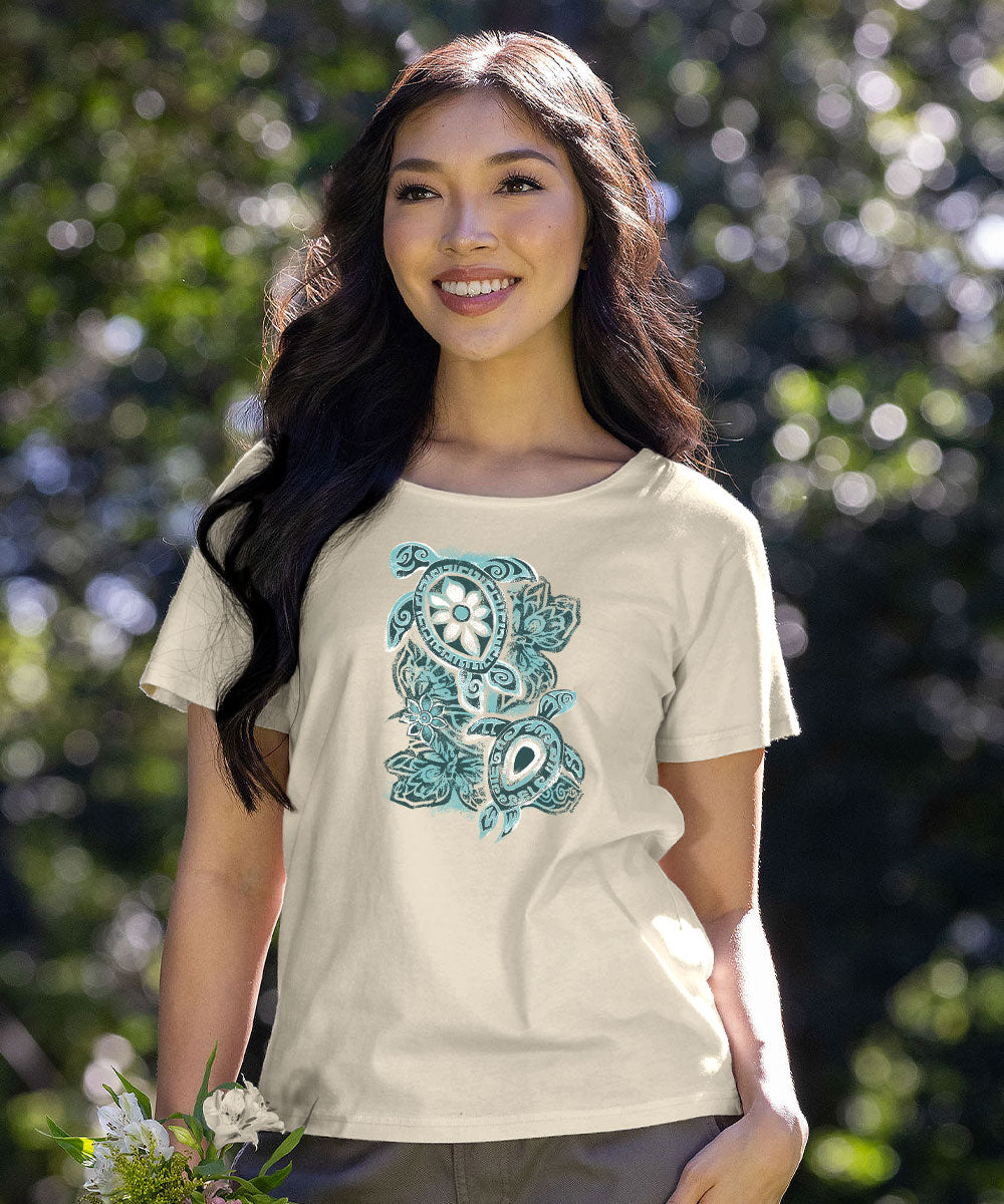 A woman wearing a cream-colored t-shirt with a teal and white tropical graphic print featuring a turtle, palm leaves, and a sun, set against a blurred green outdoor background. View 2