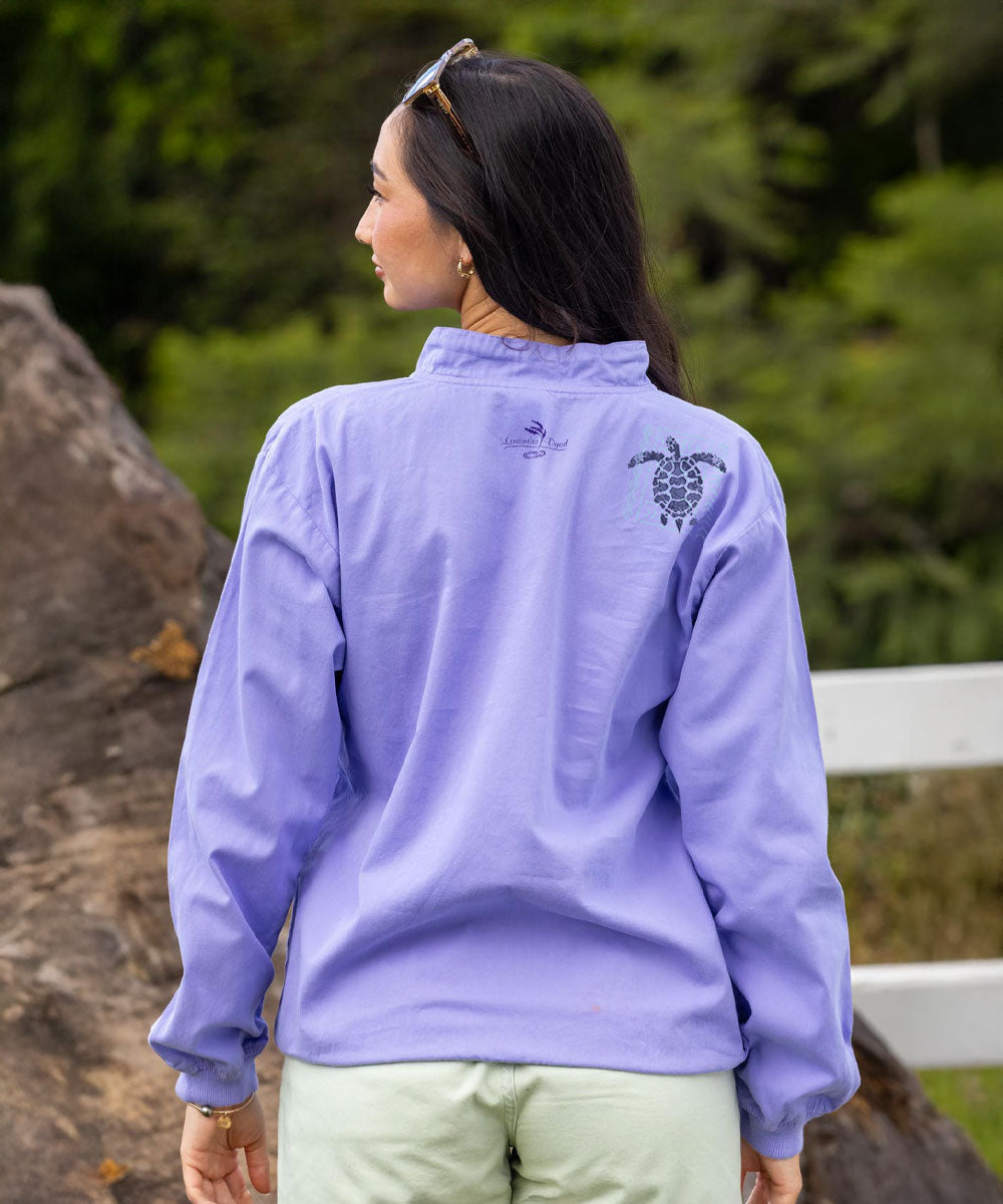 A woman viewed from behind wearing a lavender-purple lightweight windbreaker jacket with a black sea turtle graphic on the upper back, paired with light green pants, set against a blurred outdoor background of green foliage and rocks. View 3