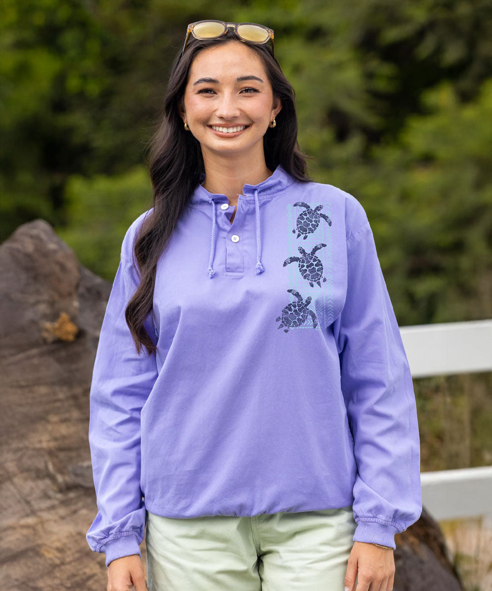 A woman wearing a lavender long-sleeve pullover top with three black turtle graphics on the left side of the chest, paired with light green pants, standing outdoors with a blurred green background. View 2